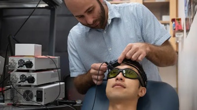 Caltech and the Keck School of Medicine of USC have developed a new laser-based headset device that can noninvasively measure blood flow and volume. Caltech postdoctoral scholar Simon Mahler is shown placing the device onto graduate student Yu Xi Huang. Mahler and Huang are co-lead authors of a paper describing the new device in the journal Biomedical Optics Express.