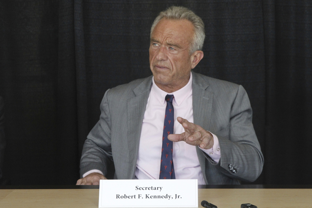 Older man in gray suit speaking at a press conference with serious expression