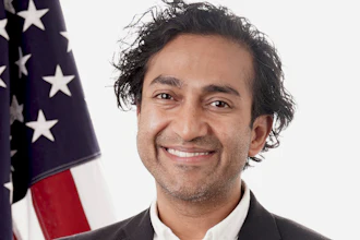 Professional headshot of a man in dark blazer and white shirt smiling in front of American flag