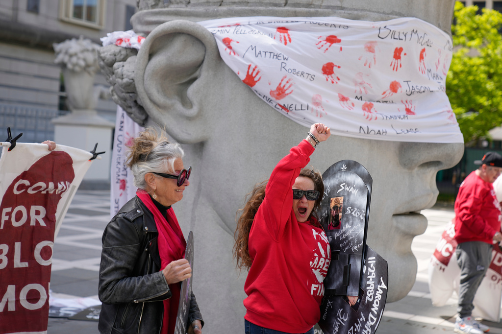 Activists in red clothing raise fists beside a large stone head sculpture wrapped with handprint-covered bandage and protest signage.