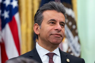 Professional man in dark suit and burgundy tie standing before American flag in formal government setting