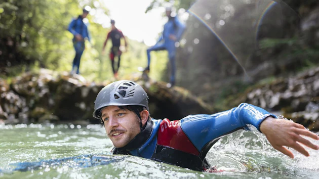 Man canyoning in turquoise river wearing helmet and blue harness with group in background