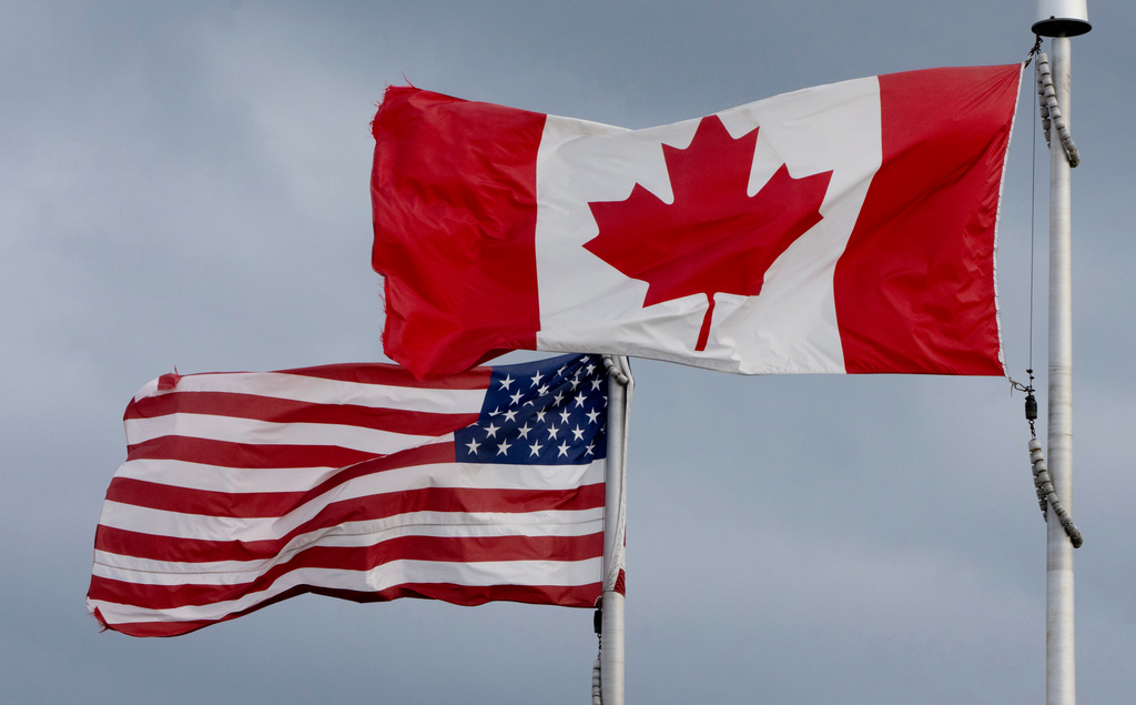 Canadian and American flags flying side by side on white flagpoles against a cloudy sky