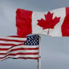 Canadian and American flags flying side by side on white flagpoles against a cloudy sky