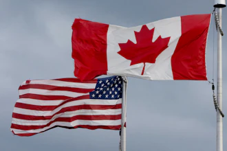 Canadian and American flags flying side by side on white flagpoles against a cloudy sky
