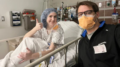 Two healthcare workers smiling in a hospital room, one wearing surgical cap and gown, the other in black scrubs with yellow mask and ID badge