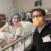 Two healthcare workers smiling in a hospital room, one wearing surgical cap and gown, the other in black scrubs with yellow mask and ID badge