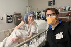 Two healthcare workers smiling in a hospital room, one wearing surgical cap and gown, the other in black scrubs with yellow mask and ID badge