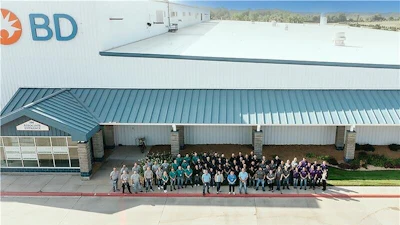 Large group of BD company employees standing together outside a modern industrial facility with white and teal metal roofing.