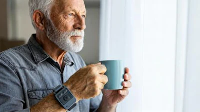 Senior man with gray beard holding a teal mug while looking out a window