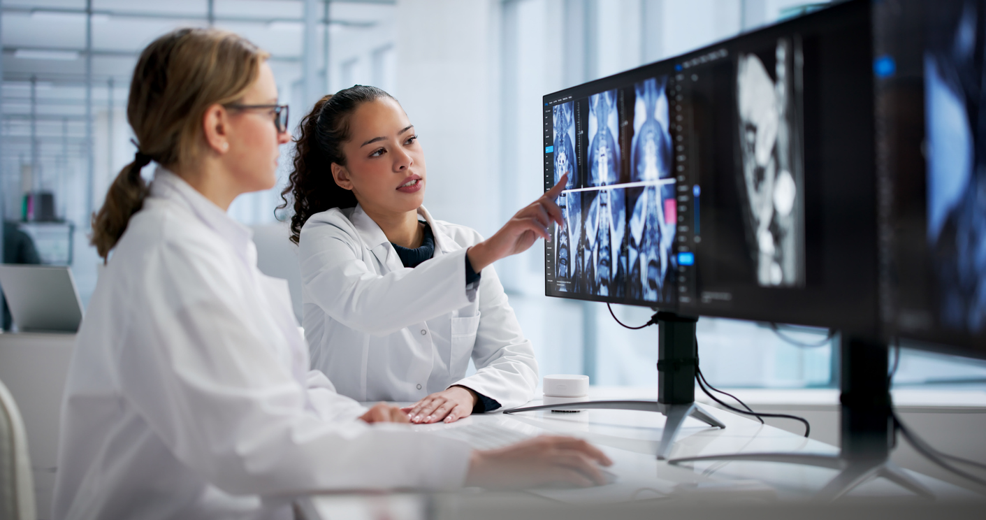 Two medical professionals in white coats review CT or MRI scans on a computer monitor in a modern clinical office setting.