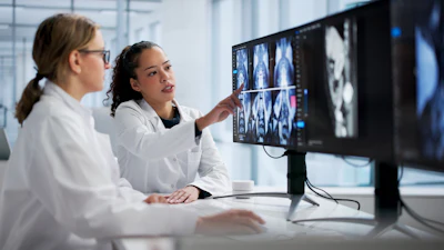 Two medical professionals in white coats review CT or MRI scans on a computer monitor in a modern clinical office setting.