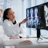 Two medical professionals in white coats review CT or MRI scans on a computer monitor in a modern clinical office setting.
