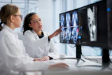 Two medical professionals in white coats review CT or MRI scans on a computer monitor in a modern clinical office setting.