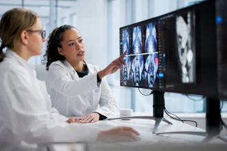 Two medical professionals in white coats review CT or MRI scans on a computer monitor in a modern clinical office setting.