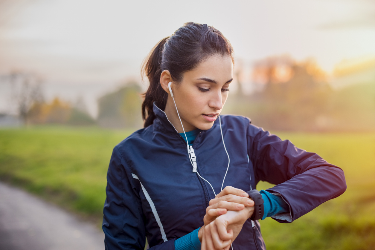 Woman jogging outdoors wearing earbuds and checking smartwatch during sunset run