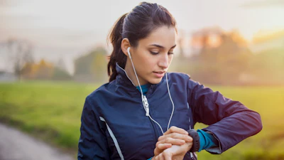 Woman jogging outdoors wearing earbuds and checking smartwatch during sunset run