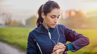 Woman jogging outdoors wearing earbuds and checking smartwatch during sunset run