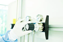 Gloved hand holding a brass gas regulator valve with pressure gauges and black control knob in laboratory setting