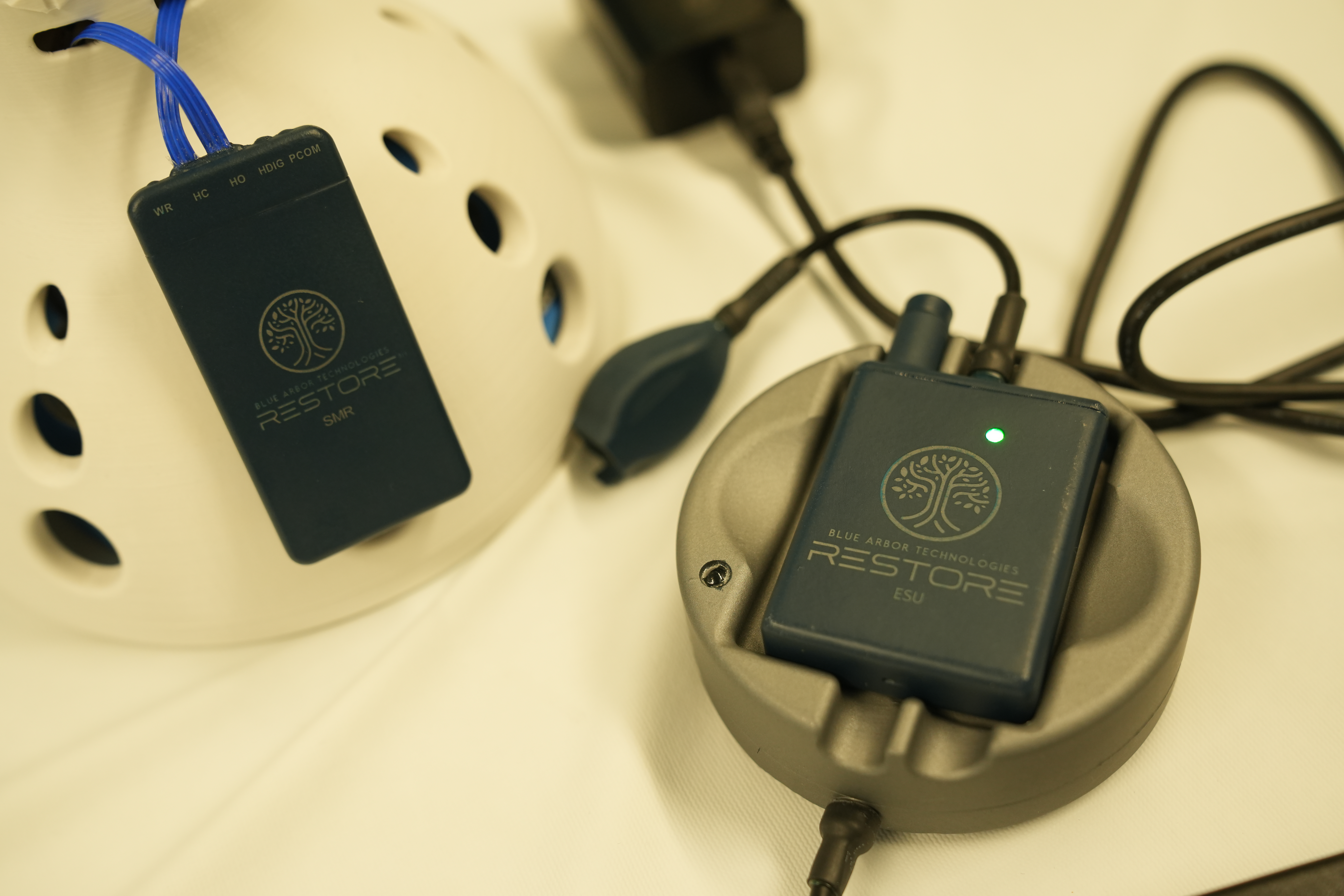 Two black Restore portable power banks with tree logo displayed on cream-colored charging dock with blue and black cables