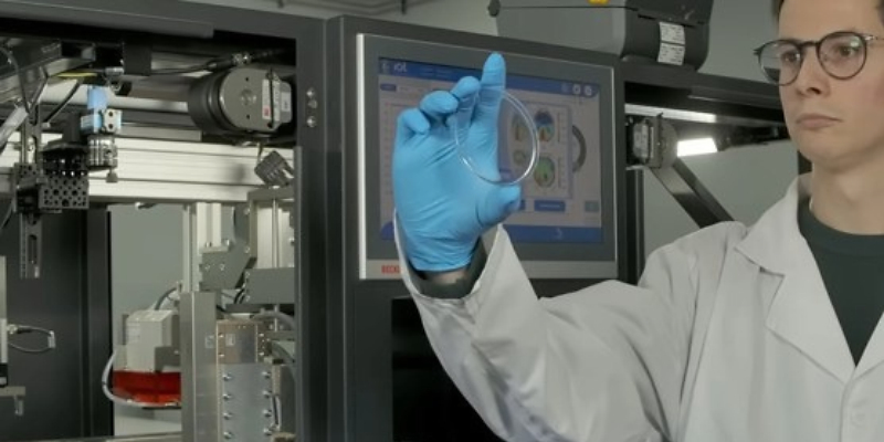 Scientist in lab coat and blue gloves examining a transparent sample near computer monitor displaying technical data