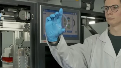 Scientist in lab coat and blue gloves examining a transparent sample near computer monitor displaying technical data