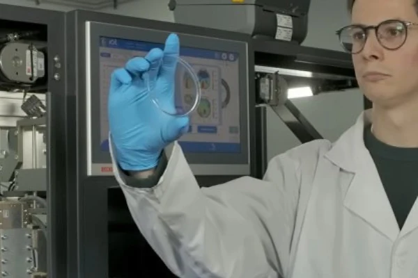 Scientist in lab coat and blue gloves examining a transparent sample near computer monitor displaying technical data