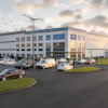 Modern industrial warehouse facility with blue and white facade, parking lot with vehicles, and landscaped grounds at dusk