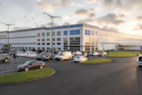 Modern industrial warehouse facility with blue and white facade, parking lot with vehicles, and landscaped grounds at dusk