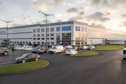 Modern industrial warehouse facility with blue and white facade, parking lot with vehicles, and landscaped grounds at dusk