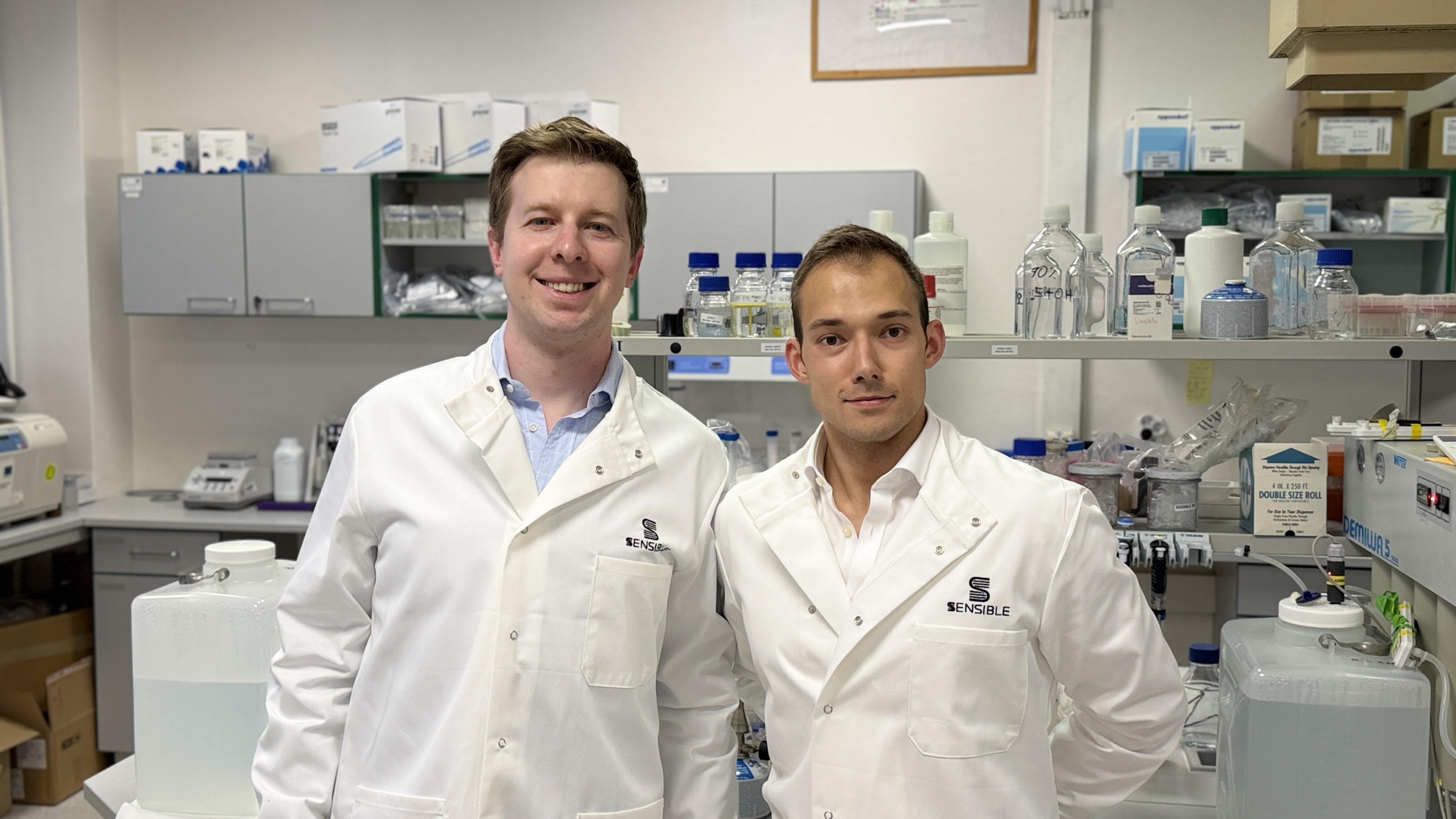 Two scientists in white lab coats smiling in a modern laboratory with equipment and supplies in the background