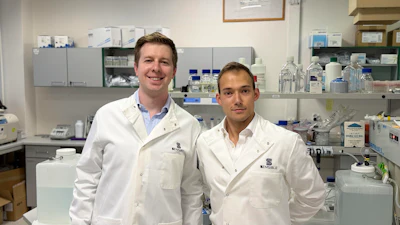 Two scientists in white lab coats smiling in a modern laboratory with equipment and supplies in the background