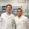 Two scientists in white lab coats smiling in a modern laboratory with equipment and supplies in the background