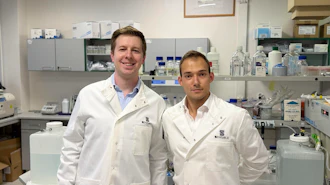 Two scientists in white lab coats smiling in a modern laboratory with equipment and supplies in the background