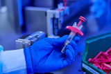 Gloved hand holding a syringe with pink plunger in laboratory setting