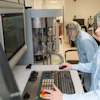 Two laboratory technicians in hairnets operating advanced analytical equipment in a clinical facility.