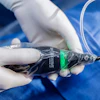Gloved hands holding a black medical device with green indicator light during clinical procedure