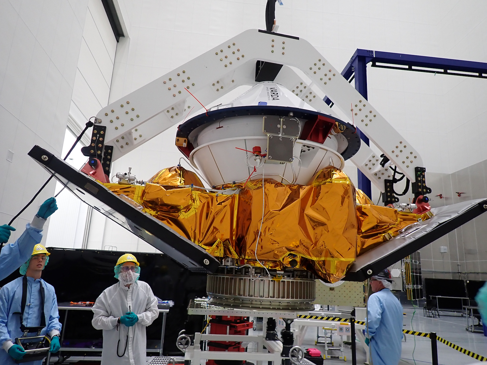 Spacecraft module with golden thermal blankets suspended in assembly facility with technicians and overhead support structure