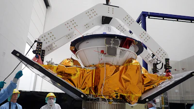 Spacecraft module with golden thermal blankets suspended in assembly facility with technicians and overhead support structure