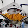 Spacecraft module with golden thermal blankets suspended in assembly facility with technicians and overhead support structure