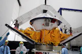 Spacecraft module with golden thermal blankets suspended in assembly facility with technicians and overhead support structure