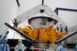 Spacecraft module with golden thermal blankets suspended in assembly facility with technicians and overhead support structure